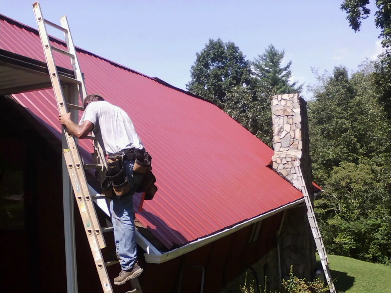 Roofer on a ladder beside a red metal roof and stone chimney — Soffit & Fascia Repair by Roofing Brothers in Leasburg